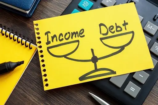 A drawing on a notebook of scales with ‘Income’ on one side and ‘Debt’ on the other placed on top of desk background.