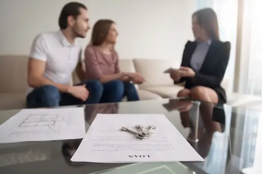 A photo of keys, loan documents and house blueprints in focus in the foreground, with a couple and a lender out of focus in the background.