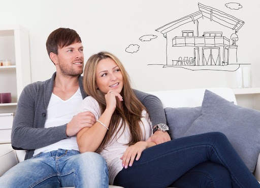A couple sitting on a couch with a thought bubble in the shape of a house drawn over their heads.