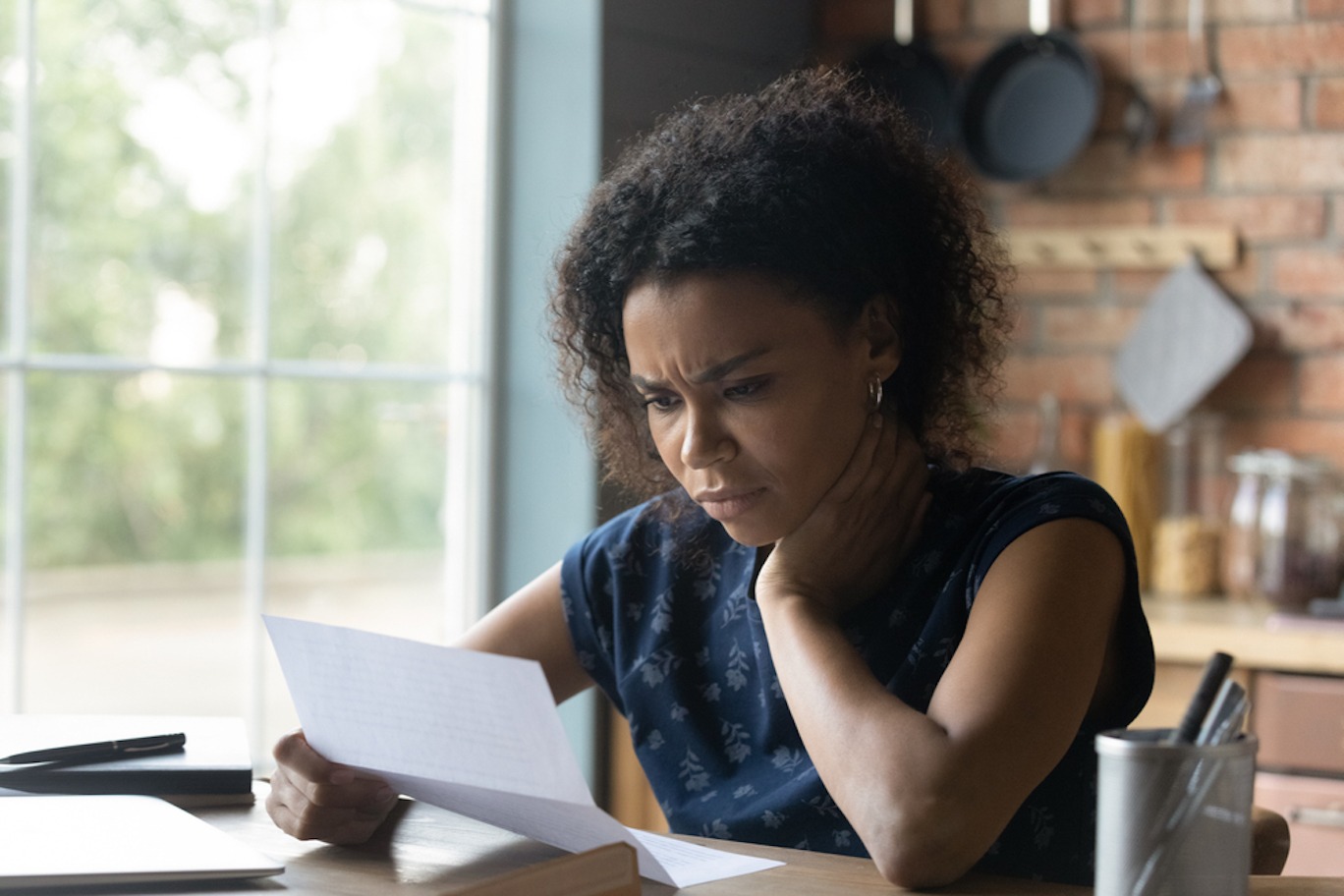 Young Black woman reading a letter with a distressed expression on her face while sitting in her kitchen.