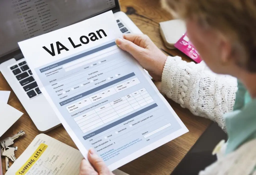 Woman holding a VA loan application while sitting at a desk cluttered with papers and her laptop.