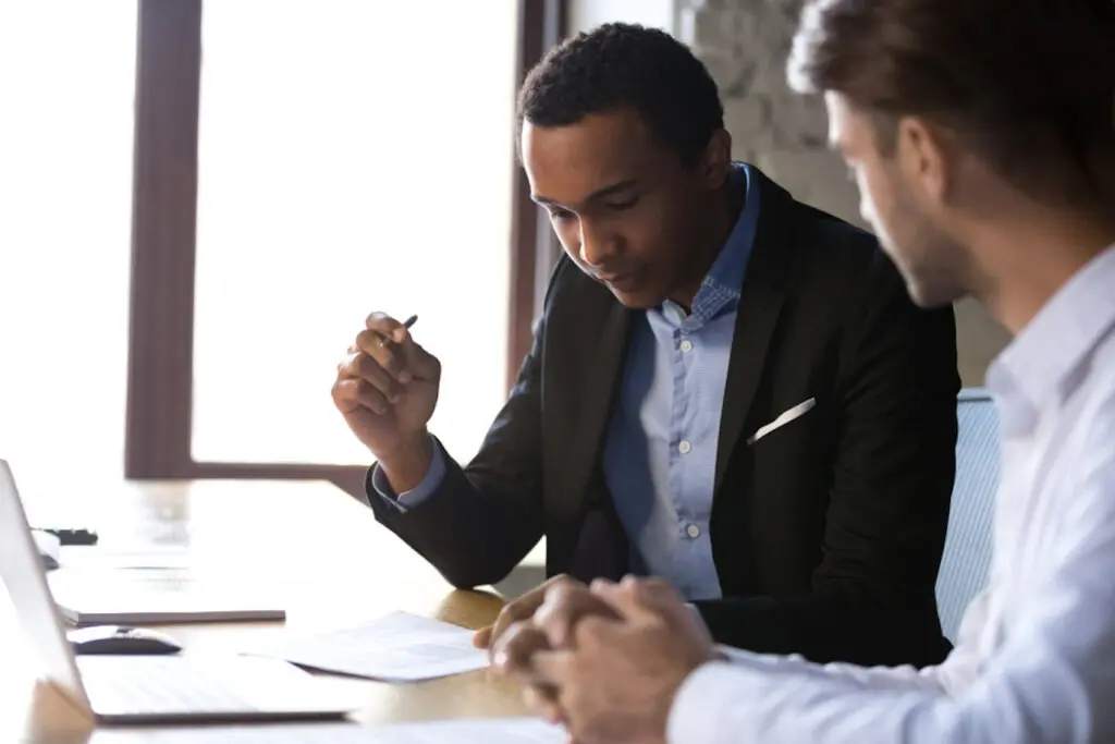 Image of a Black man reviewing documents sitting next to a white man at a table with a laptop on it.