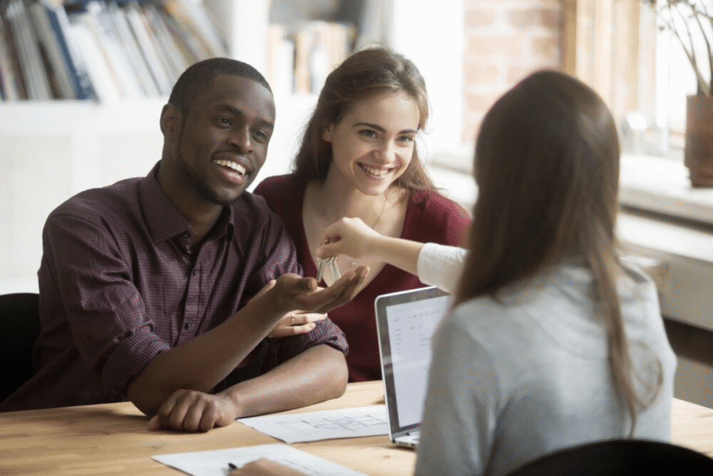 Smiling couple receives house keys from a loan officer at a desk with a laptop and papers after closing on a no doc mortgage for their new home.