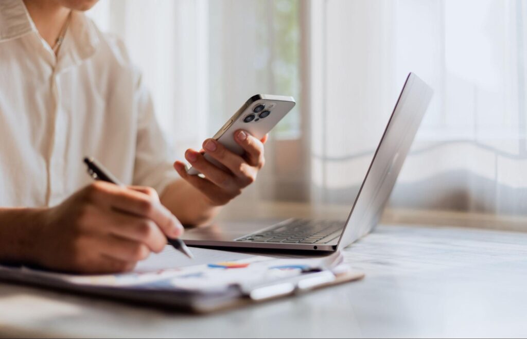 Image of a man using his phone and writing something down with a laptop in front of him.