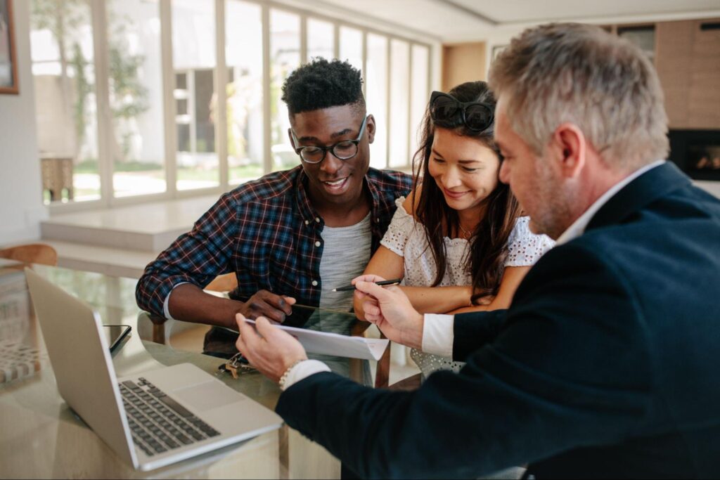 A young couple sitting at a table excitedly looks over a contract while being handed a pen by an older real estate agent.