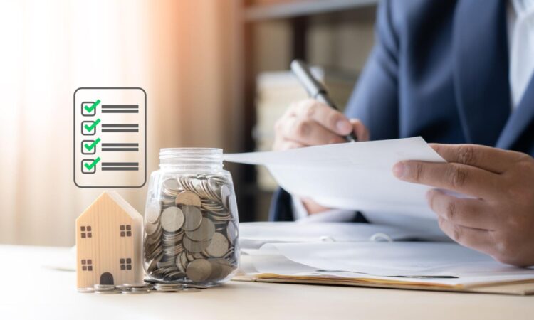 A person signs a document while in the foreground there is a checklist graphic, a small model home, and a jar filled with coins.