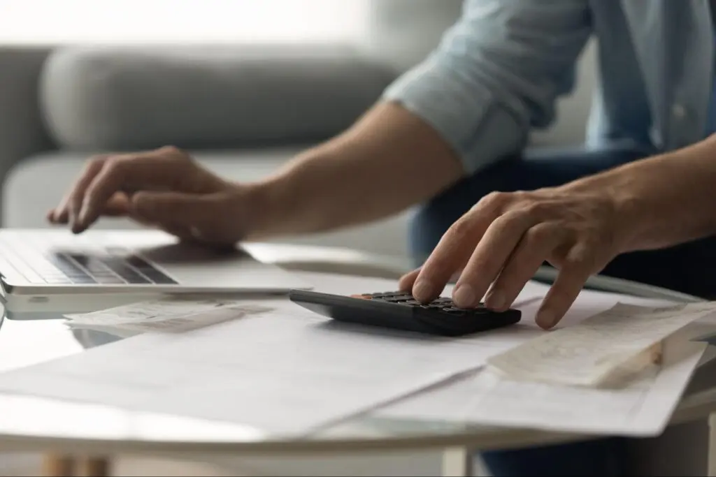 Close up of man’s hands as he types on a keyboard and calculator with papers spread out in front of him on a table.
