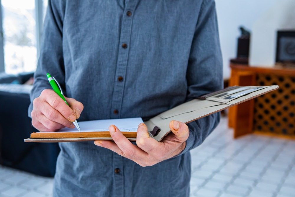 Close up of a man holding a folder with a clipboard and writing on a notepad while standing in a living room.