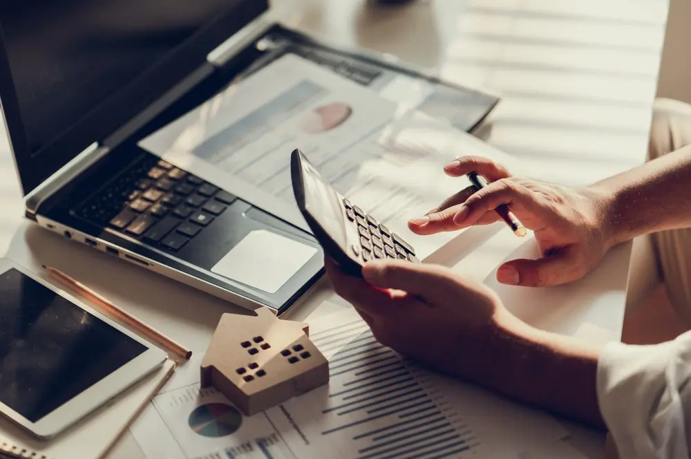 A person types on a calculator over a desk that has an open laptop on it, a small cutout of a house, and several charts and graphs.