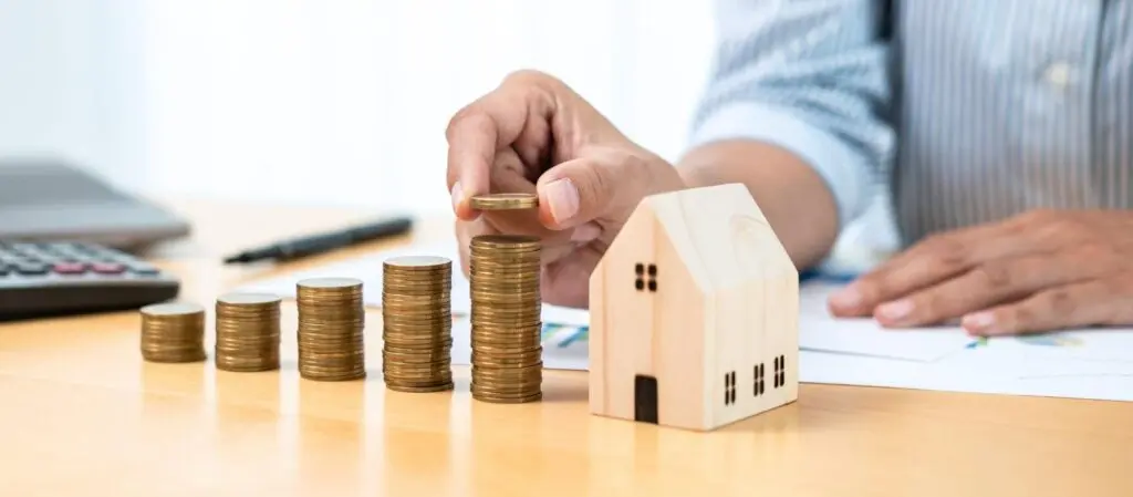 A small model of a home sits next to gradually rising stacks of coins and a person is placing another coin on top of the tallest stack