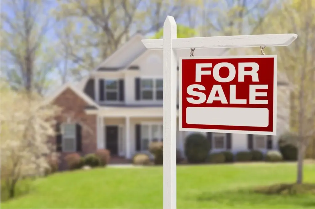 Image of a two story house surrounded by trees with a for-sale sign out front.