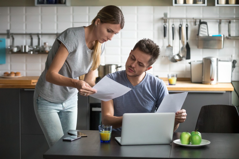 Young couple at kitchen table reviewing their finances. Woman is holding papers while the man is using the laptop in front of him.