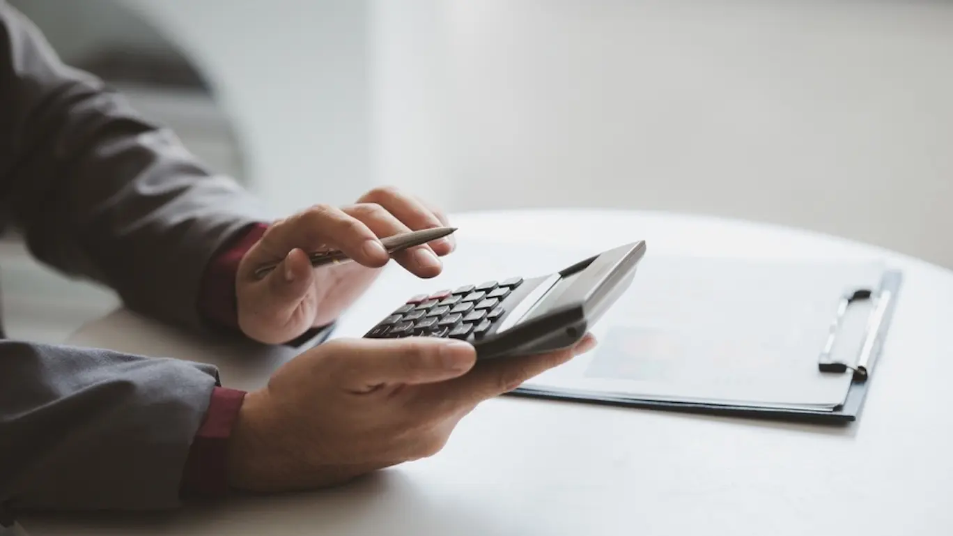 The hands of a person holding a calculator over a table that has a clipboard with a document attached to it.
