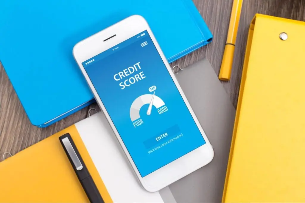 A smartphone lying on top of blue and orange binders with a blue screen that has a credit score meter with the needle leaning towards “Good”.