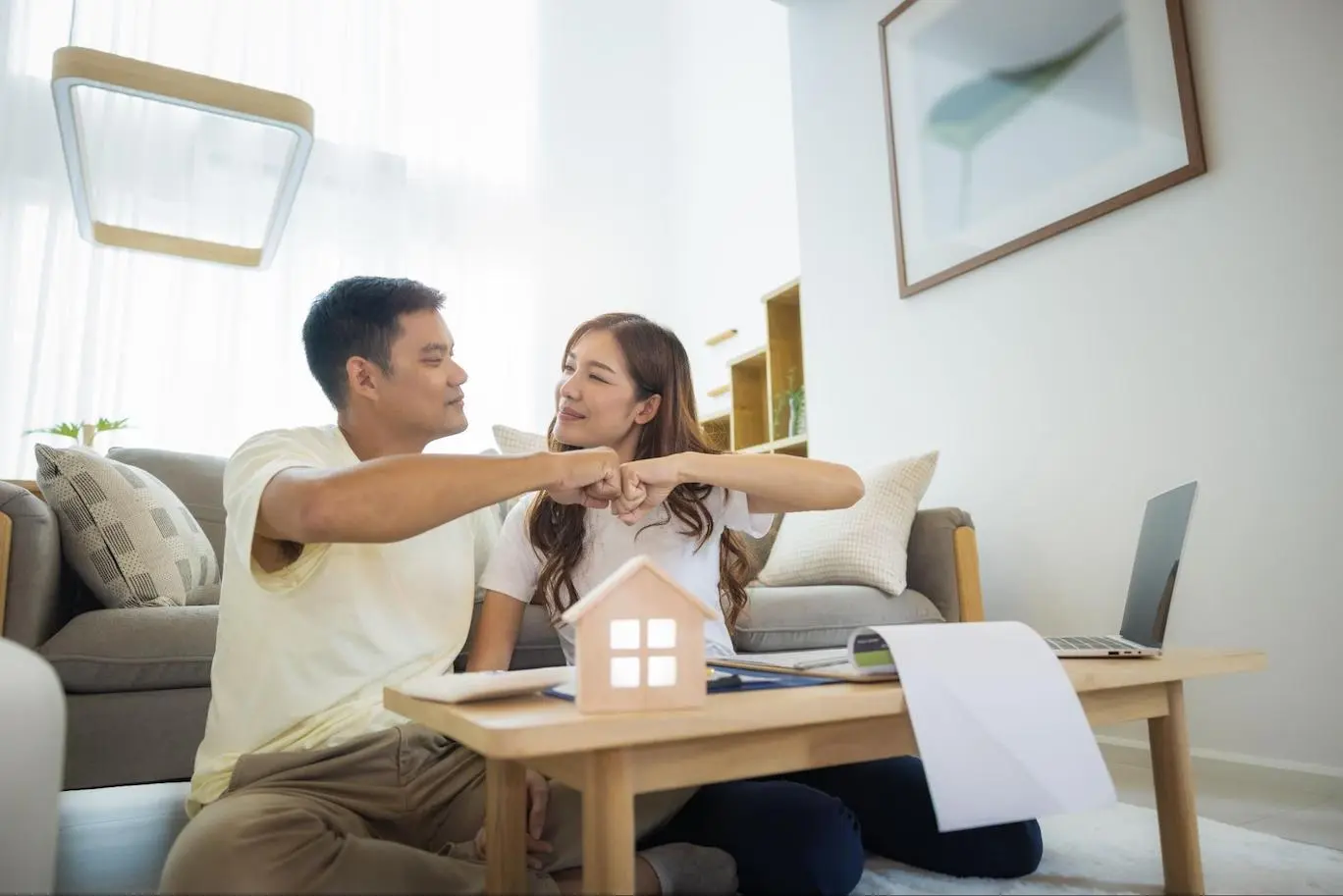 An Asian man and woman sitting in front of a couch and behind a desk cluttered with paperwork, an open laptop, and a small home model smile at each other and bump fists.
