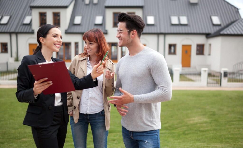 A young couple stands beside a smiling real estate agent who’s holding a red clipboard and gesturing with a pen in her other hand.