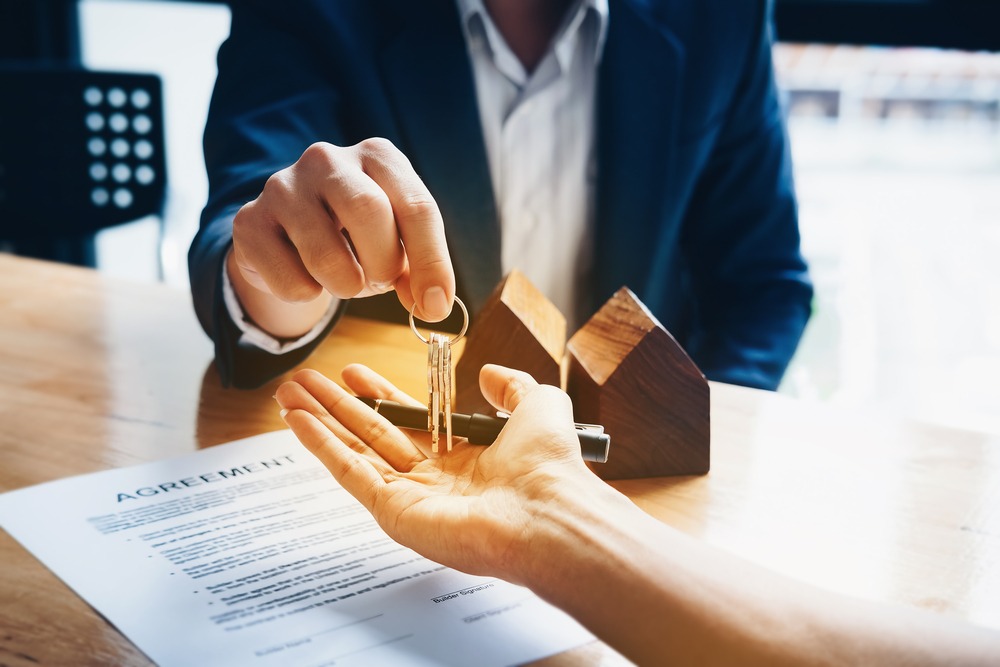 One person in a dark suit hands another person the keys to a home over a table that has a contract sitting on it as well as two small wooden home models.