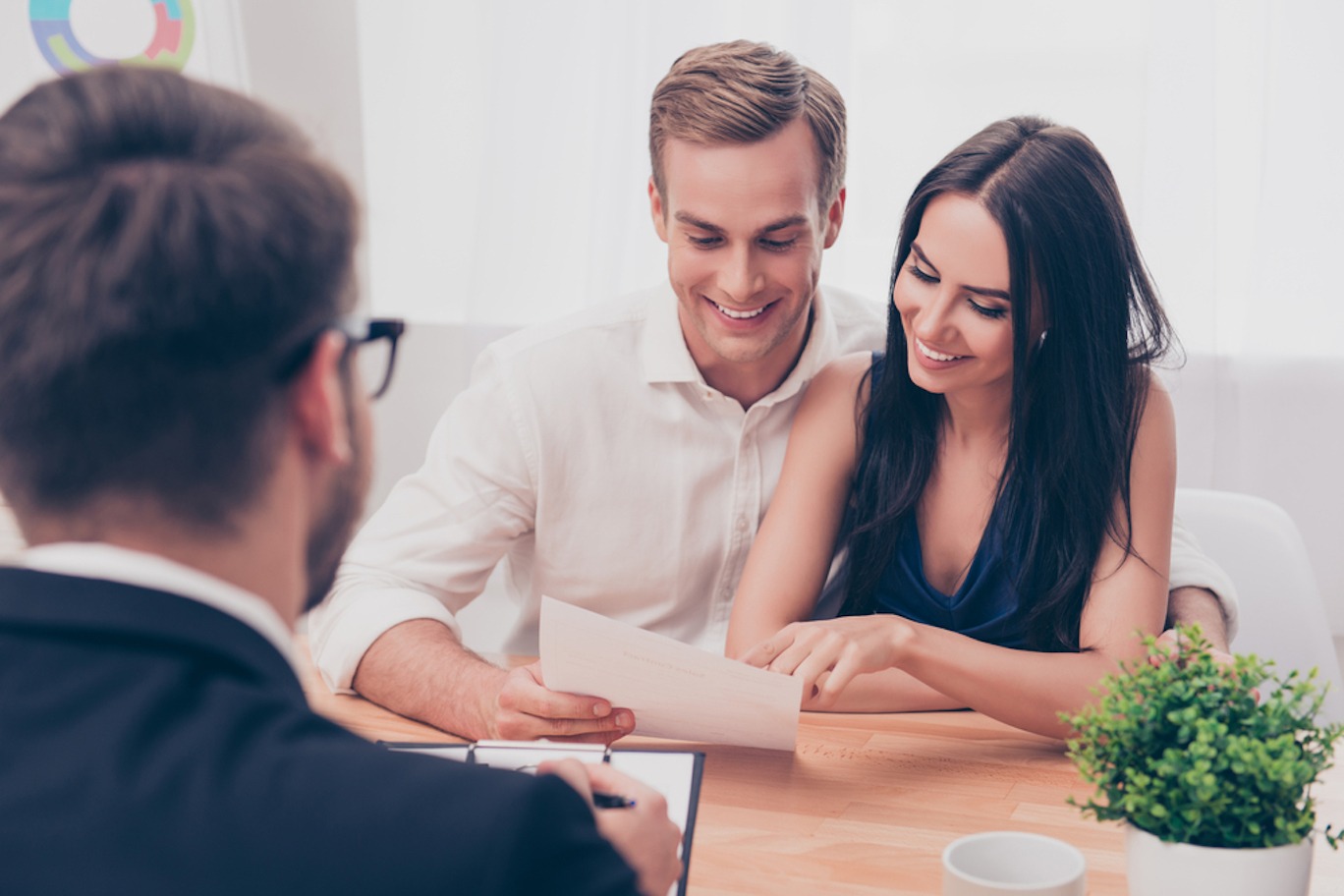 A smiling man and woman sit opposite from their realtor and look over a document together.