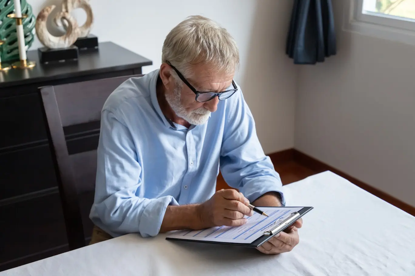 An older man in a light blue collared shirt and glasses sits at a table and reads over a document attached to a clipboard.