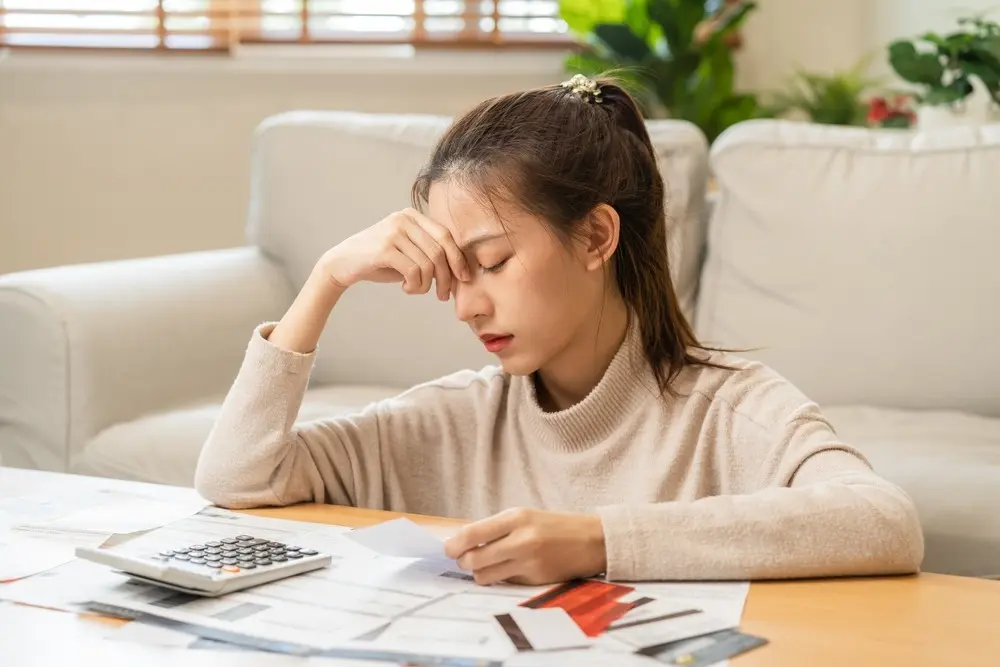 A stressed out Asian woman sits in front of a coffee table cluttered with a calculator, financial documents, and credit cards.