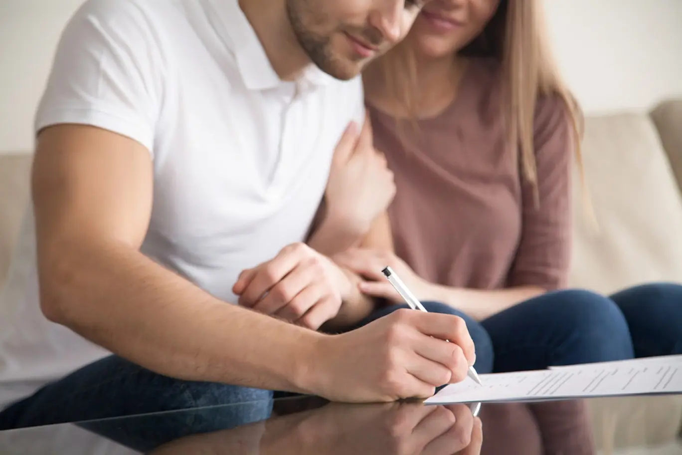 A man in a white polo signs a piece of paper while a woman holds onto his arm and watches.