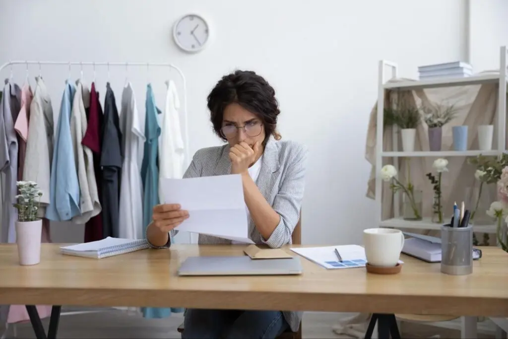 A white woman sits at a desk in her shop and reviews a document with a concerned look on her face.