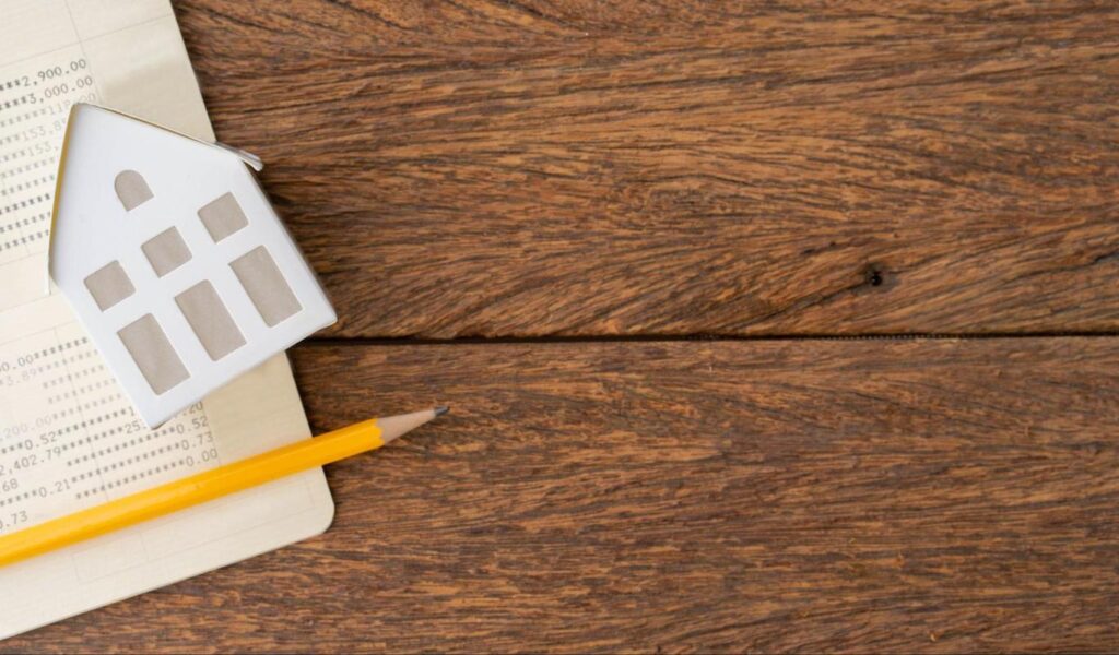 A small model home sits on top of a paper bank statement and a pencil.
