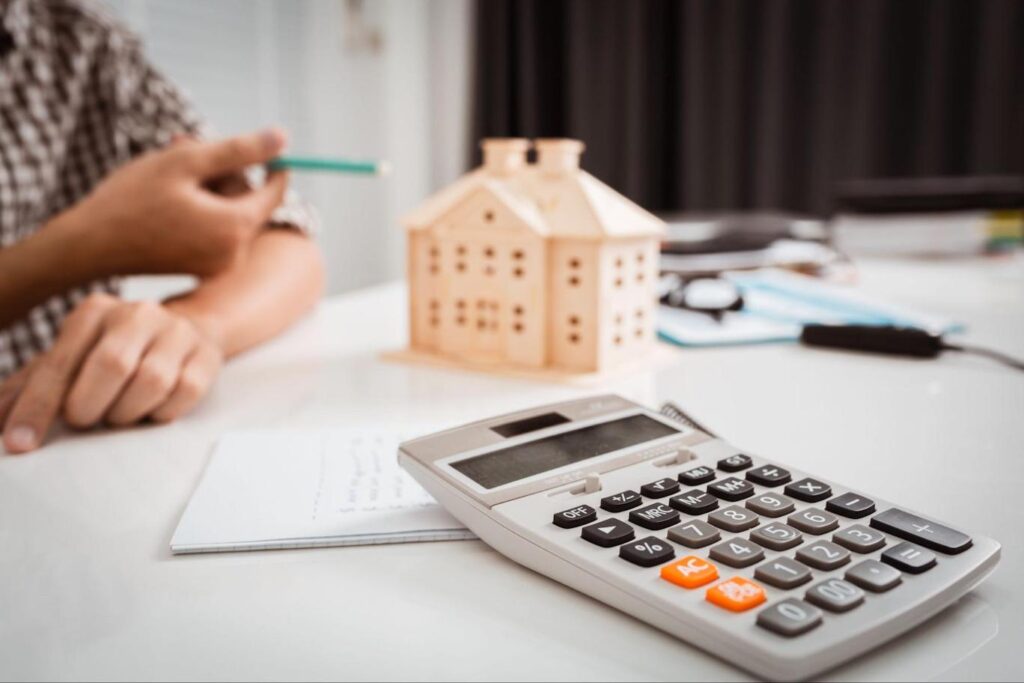 A hand gestures to a small model of a house sitting on a cluttered table while a calculator sits in the foreground.