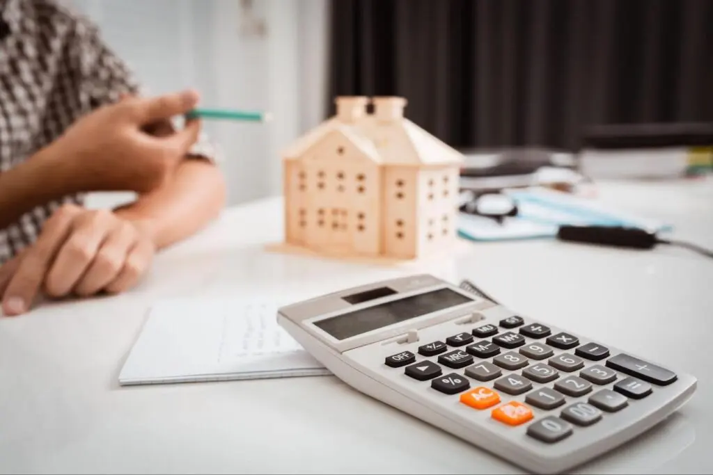 A hand gestures to a small model of a house sitting on a cluttered table while a calculator sits in the foreground.