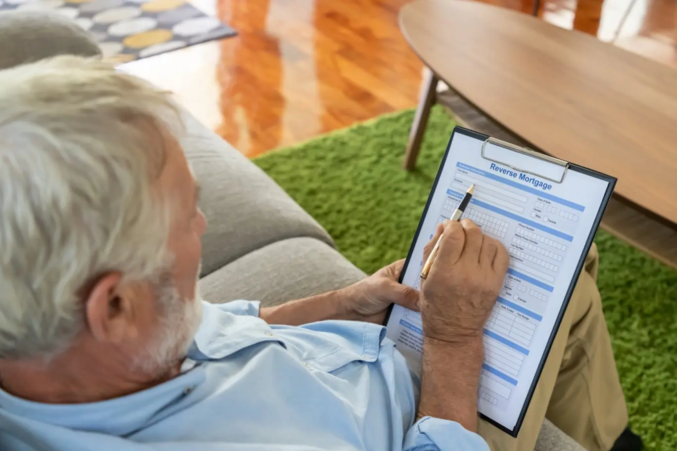 An older man with white hair and a light blue collared shirt sits on a couch and reviews a reverse mortgage application with pen in hand.