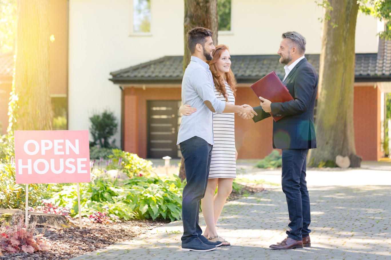 A real estate agent in a dark suit shakes hands with a young couple while standing outside beside an open house sign.