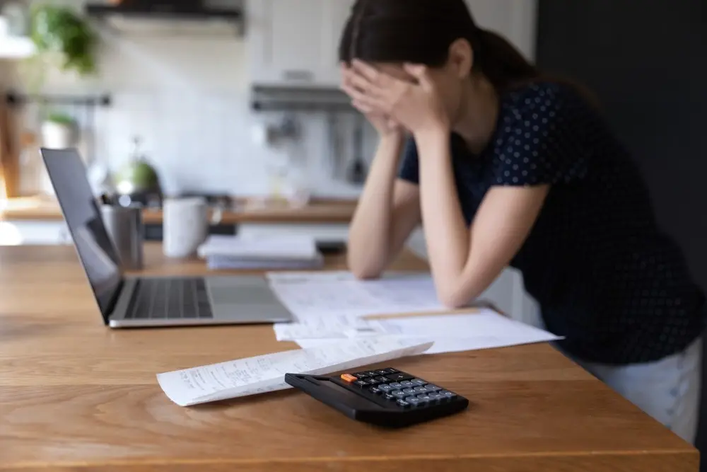 A stressed woman sits at a desk with her head in her hands and an open laptop, calculator, and various documents in front of her.