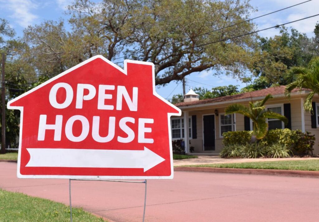 A bright red open house sign is posted on the grass in front of a home.