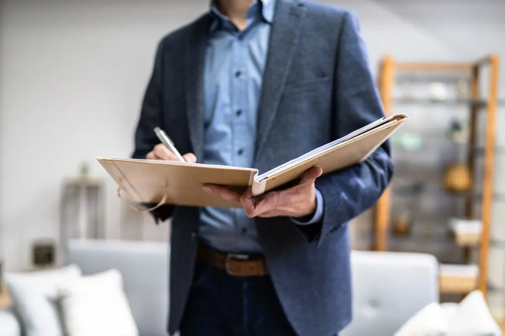 A home appraiser in a blue button up shirt and blazer stands in the room of a home and writes on a clipboard.
