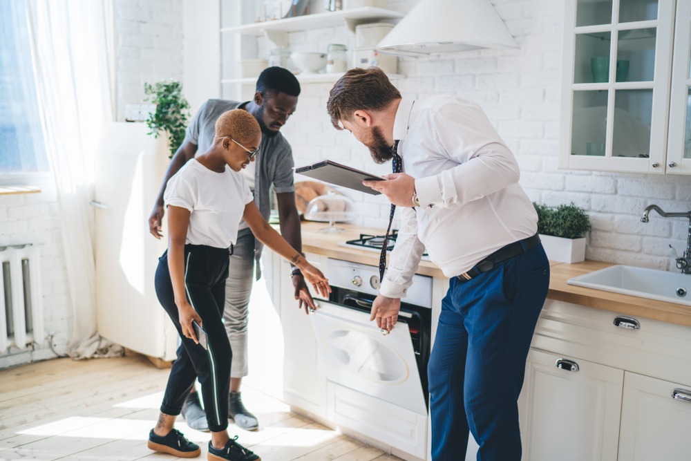 A real estate agent in a white shirt opens the oven in a home while giving a tour to a young black couple.