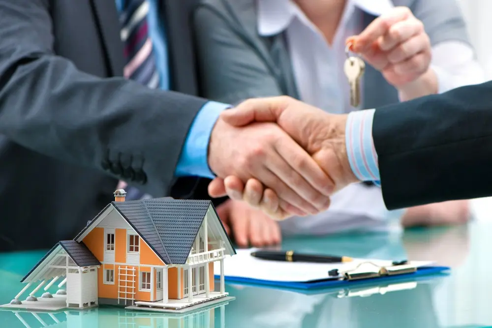 Two people wearing suits shake hands over a table that has a clipboard and an orange model home on it while another hand in the background holds a set of keys.