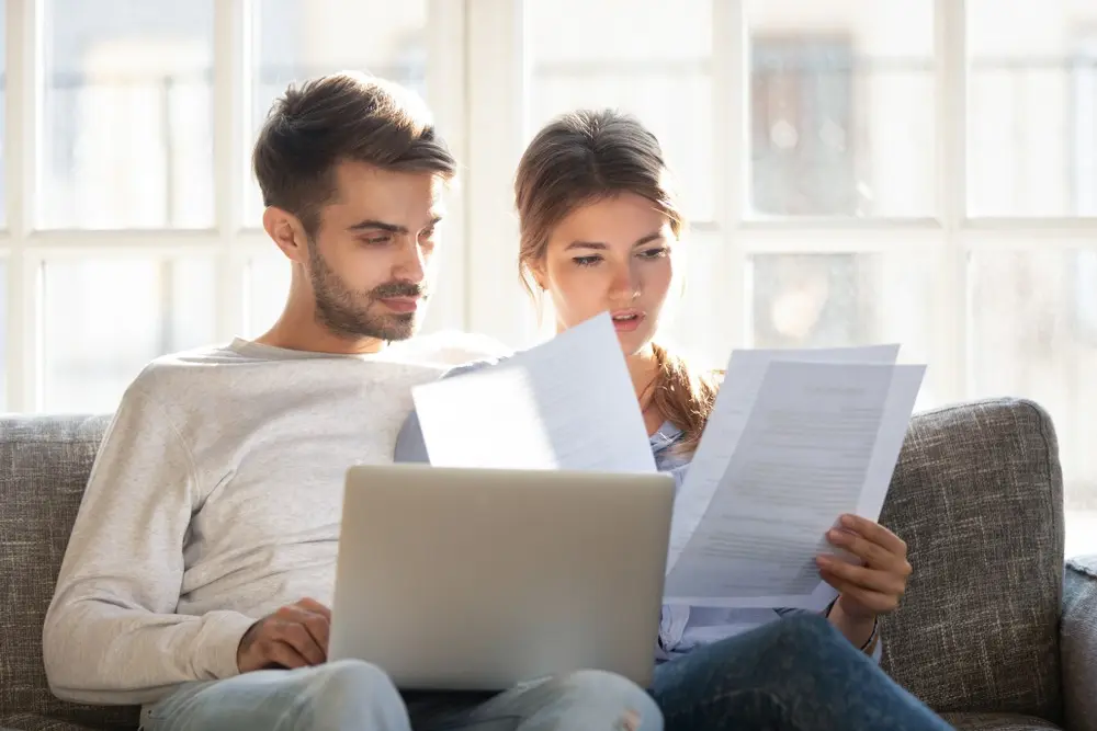 A caucasian man and woman sitting on the couch and looking through various documents.
