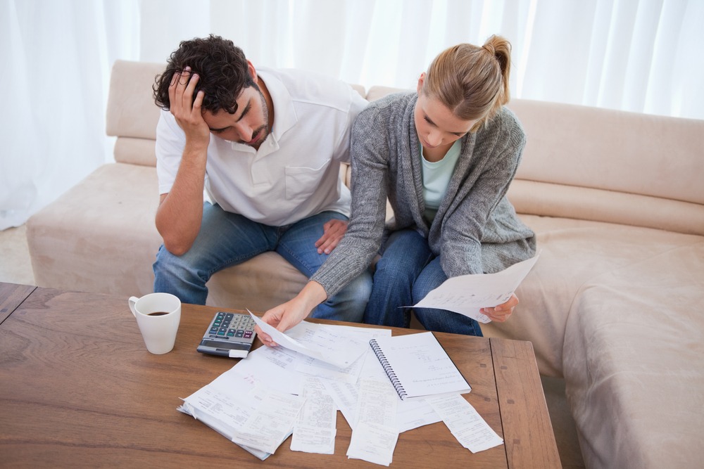 A distraught man and woman sit on the couch and review documents scattered across the coffee table.