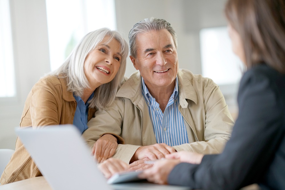 An elderly white couple smiling at their lender, who’s presenting them with a document.
