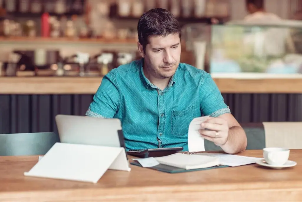 A small business owner in a teal shirt sits at a desk and reviews receipts while filing taxes.