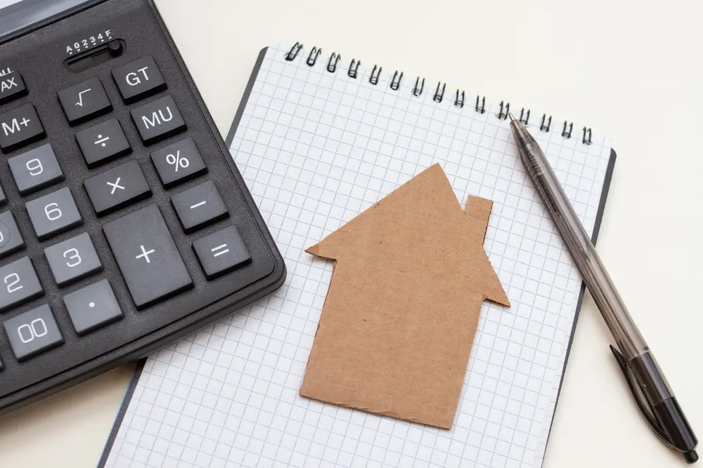 A cardboard cutout of a home sits on top of a notepad and next to a pen and calculator.
