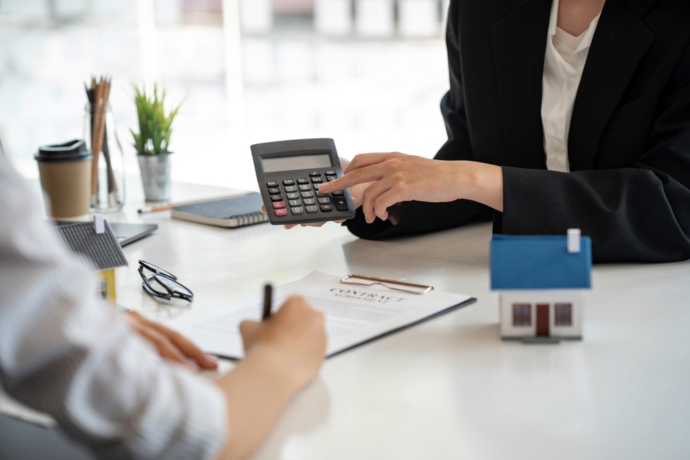 A person holds up and points to a calculator while another person signs a contract that’s lying on a table next to a model home.