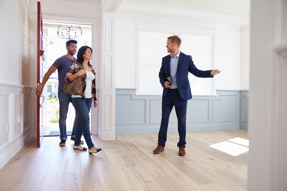 A white realtor in a blue suit welcomes a Hispanic couple into a home and prepares to show them around. 