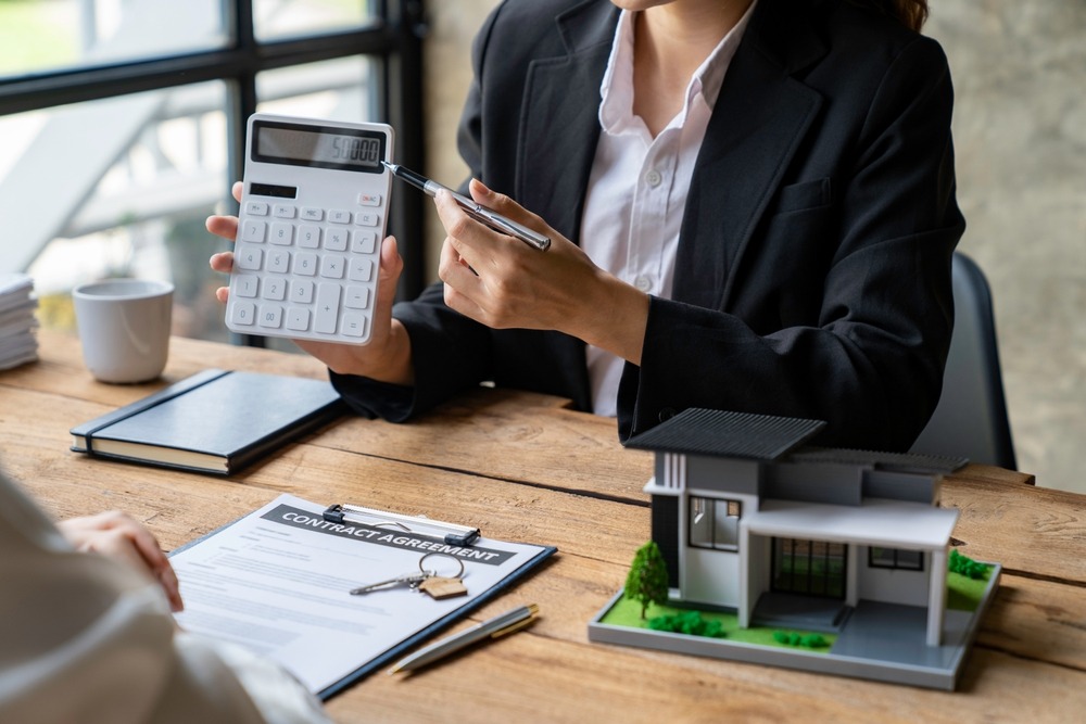 A person in a white shirt and black blazer holds up and points to a calculator that they’re holding over a desk with a contract, house keys, and model home on it.