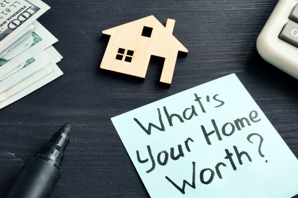 A cutout of a home lays on a dark table with cash, a calculator, a marker, and a note that says “What’s your home worth?”