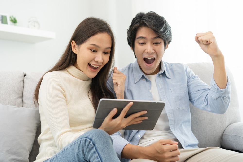 A young Asian woman and man sitting on a couch and looking at a tablet while holding their hands up in a celebratory pose. 