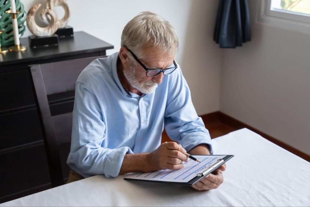 An elderly white man in a blue button up reviews a document on a clipboard.