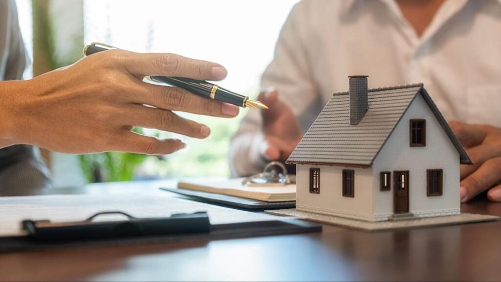 A model of a home sits on a table where two people are signing and reviewing documents and contracts.