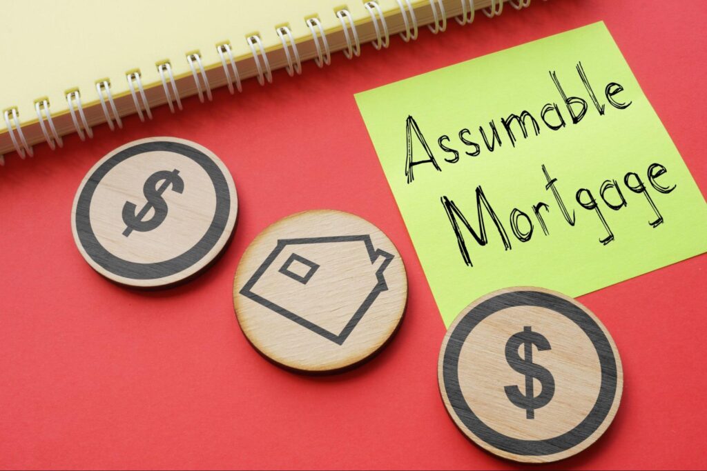 Small wooden medallions with dollar signs and an outline of a house are arranged next to the spine of a yellow notebook and a post-it note that has “Assumable Mortgage” written in pen on it.