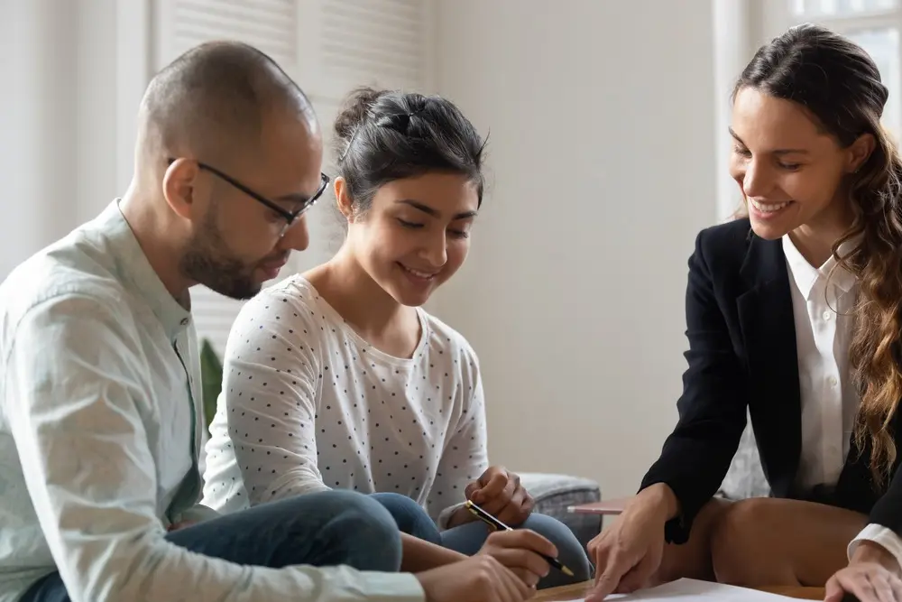 A female mortgage lender in a dark blazer points out where to sign a document for the couple sitting across from her.